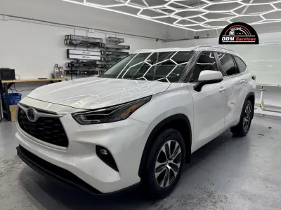Front three-quarter view of a white Toyota Highlander in a professional garage, showcasing a clean vinyl wrap finish under hexagonal LED lighting.